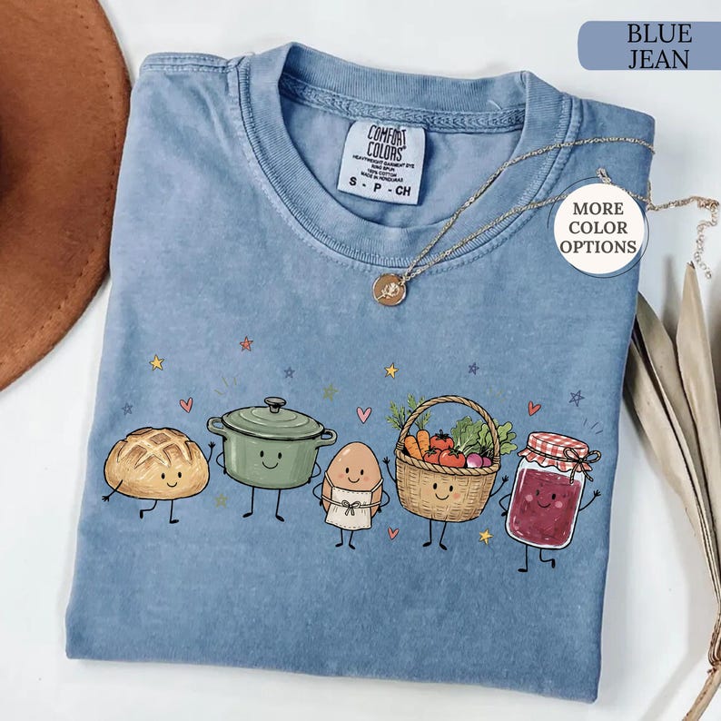 May include: A blue jean colored t-shirt featuring a graphic print of five anthropomorphic food items including a loaf of bread, a green pot, an egg wearing an apron, a basket of vegetables, and a jar of jam, accented with small stars and hearts.