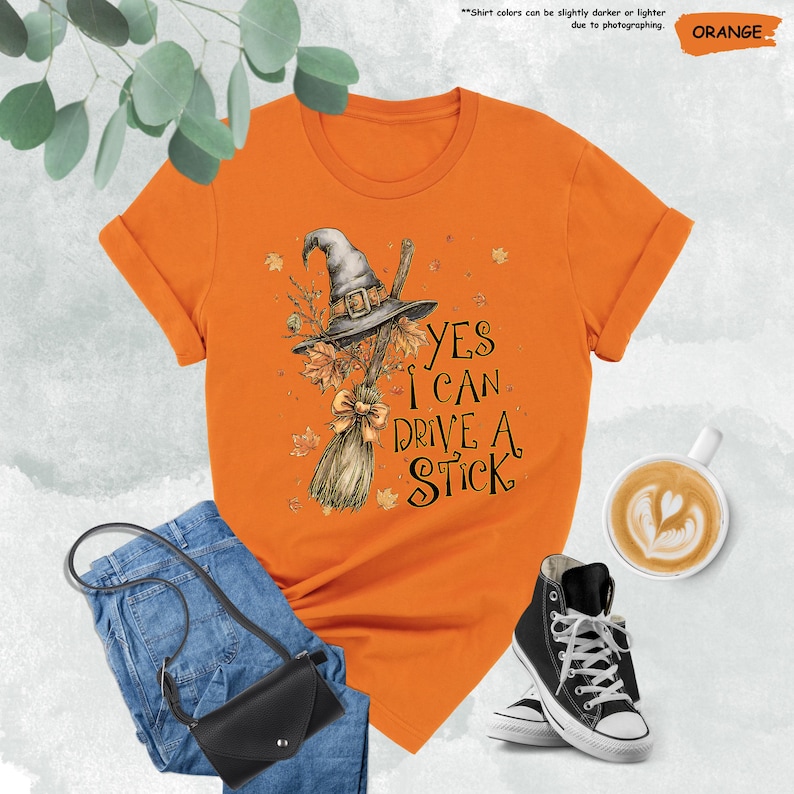 May include: An orange short-sleeved t-shirt featuring a graphic of a grey witch hat and a broomstick with autumn leaves. The text reads, "YES I CAN DRIVE A STICK." The shirt is displayed with blue jeans, a black crossbody bag, and black sneakers.