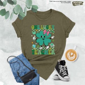 May include: A green t-shirt with a four-leaf clover graphic and the text "ONE LUCKY TEACHER". The clover has a pink bow and is holding a pencil and a globe.