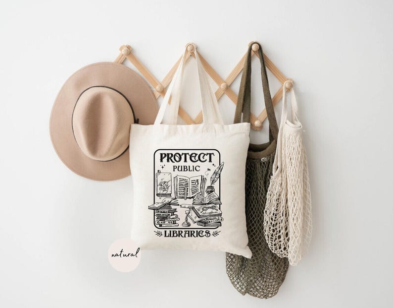 May include: A white tote bag with black text that reads "Protect Public Libraries". The bag has a graphic of a stack of books, a quill pen, and a magnifying glass. The bag is hanging on a wooden rack with a brown hat and two string bags.