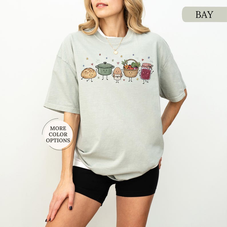 May include: A sage green short-sleeve t-shirt featuring a graphic print of five anthropomorphic food items including a loaf of bread, a green pot, an egg, a basket of vegetables, and a jar of jam, with the text "BAY" and "MORE COLOR OPTIONS".