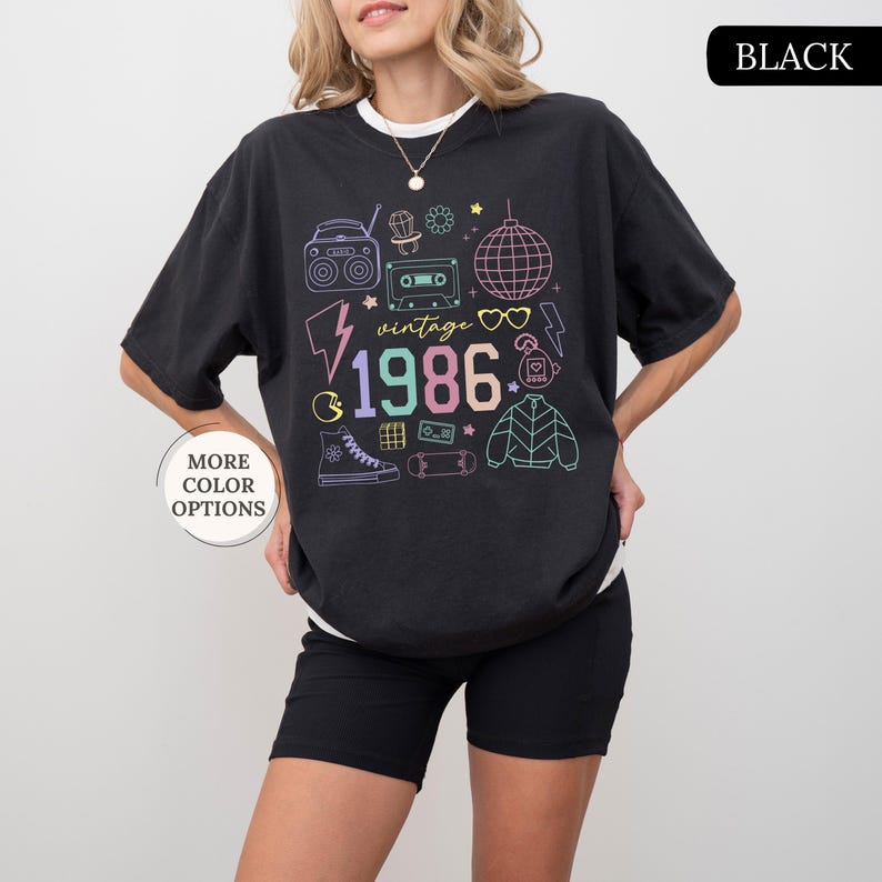 May include: Black t-shirt with a white collar featuring a colorful 1986 graphic design. The design includes a boombox, cassette tape, disco ball, lightning bolt, and other vintage icons. The shirt also has the text "vintage".