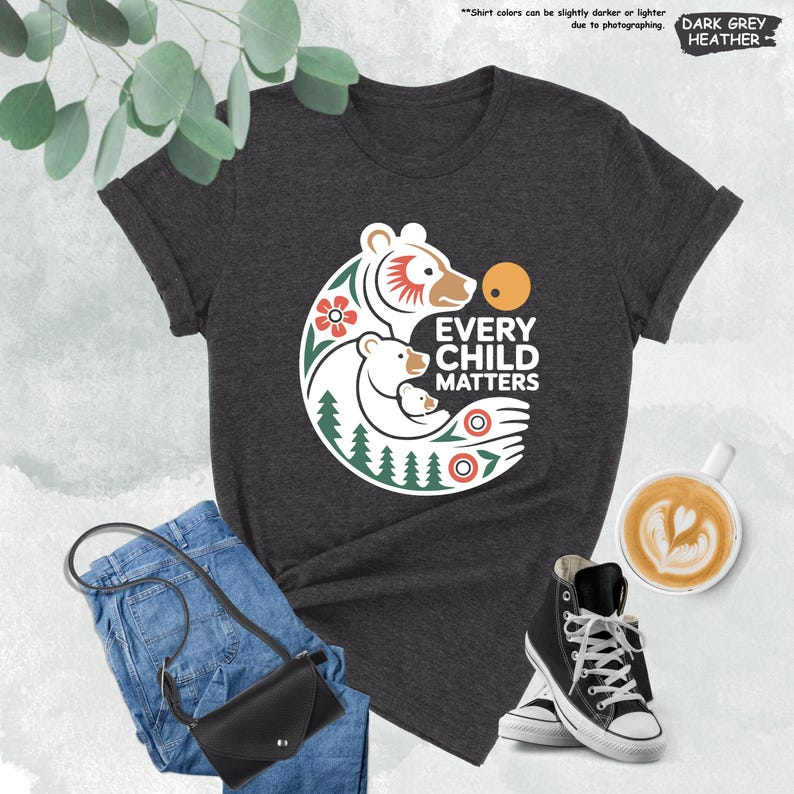 May include: Dark grey heather t-shirt with the text "EVERY CHILD MATTERS" and a graphic of a bear family. The design includes a bear, a cub, and a sun, with floral and tree elements. The shirt is paired with blue jeans, a black purse, and black high-top sneakers.