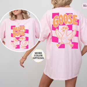 May include: A light pink t-shirt featuring a large graphic on the back with a checkered pattern in hot pink and orange, the word GOOSE in orange, and a cartoon goose wearing sunglasses. A circular inset shows the front with DUCK DUCK text.