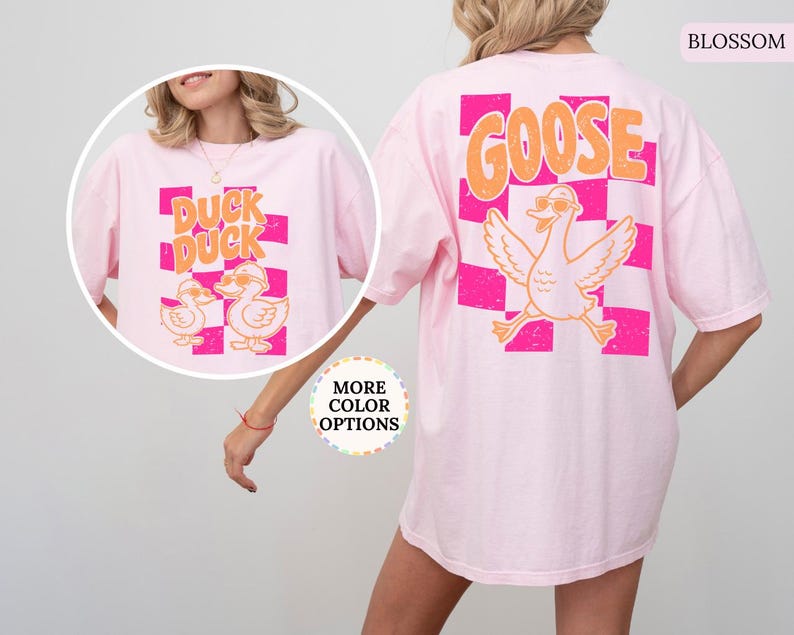 May include: A light pink t-shirt featuring a large graphic on the back with a checkered pattern in hot pink and orange, the word GOOSE in orange, and a cartoon goose wearing sunglasses. A circular inset shows the front with DUCK DUCK text.