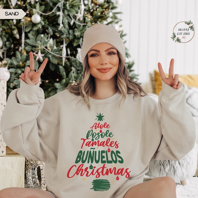 May include: A beige sweatshirt with a green and red Christmas tree design. The tree is decorated with the words "Atole", "Pozole", "Tamales", "Buñuelos", and "Christmas".