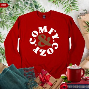 以下が含まれることがあります： 赤いスウェットシャツに、白い文字で「Comfy & Cozy」と円形に書かれています。スウェットシャツは、茶色のスカーフ、赤いリボンのかかったギフトボックス、ホットチョコレートの入ったマグカップと一緒にテーブルの上に置かれています。