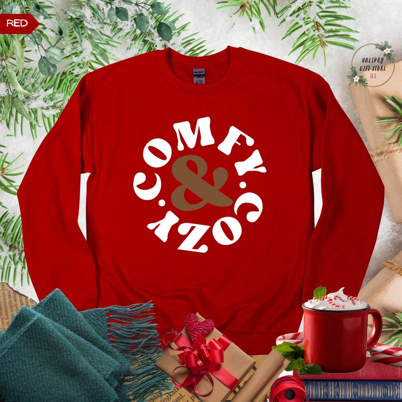 以下が含まれることがあります： 赤いスウェットシャツに、白い文字で「Comfy & Cozy」と円形に書かれています。スウェットシャツは、茶色のスカーフ、赤いリボンのかかったギフトボックス、ホットチョコレートの入ったマグカップと一緒にテーブルの上に置かれています。