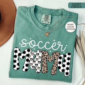 May include: Light green t-shirt with the words "soccer mama" in white script and block letters. The letters "M" are decorated with soccer balls and leopard print. The shirt has a crew neck and short sleeves. A gold necklace is draped across the shirt.