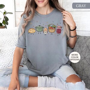 May include: A gray t-shirt featuring a graphic print of five anthropomorphic food items: a loaf of bread, a green pot, an egg, a basket of vegetables, and a jar of jam. The design includes small stars and hearts scattered above the characters.