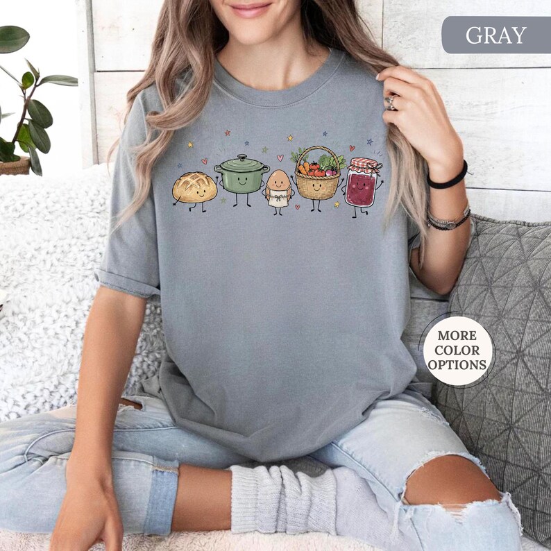May include: A gray t-shirt featuring a graphic print of five anthropomorphic food items: a loaf of bread, a green pot, an egg, a basket of vegetables, and a jar of jam. The design includes small stars and hearts scattered above the characters.