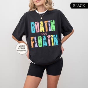 May include: Black t-shirt featuring the text "BOATIN and FLOATIN" in a distressed, multi-coloured font. The shirt is worn with black shorts. The words "MORE COLOR OPTIONS" are in a circle on the left.