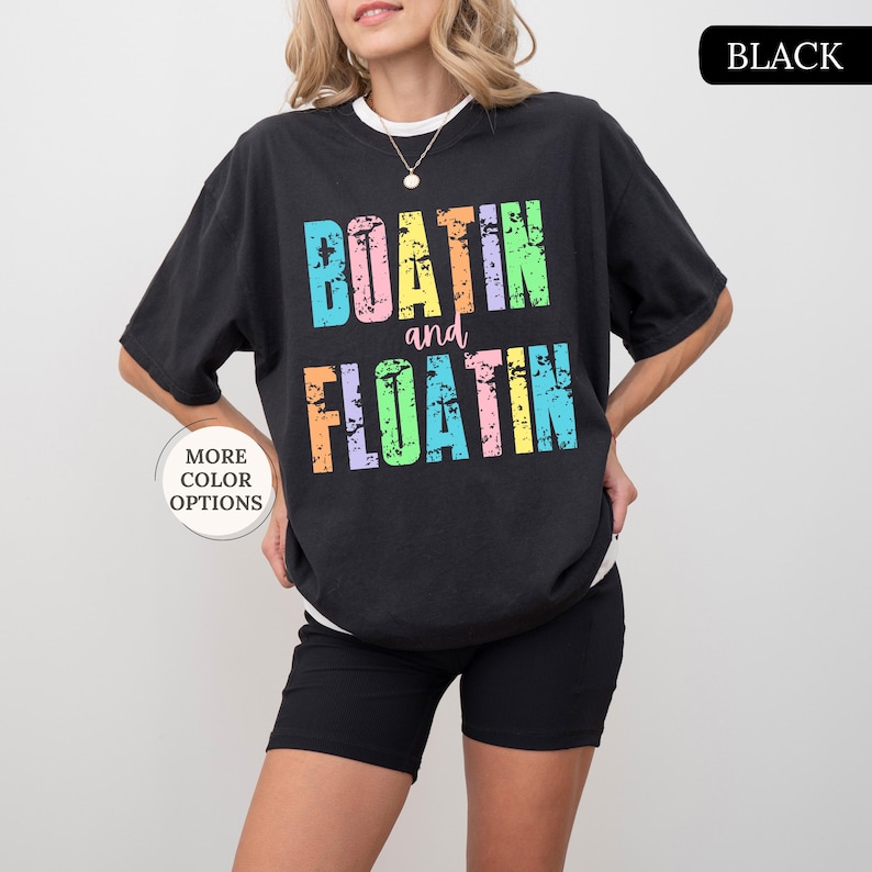 May include: Black t-shirt featuring the text "BOATIN and FLOATIN" in a distressed, multi-coloured font. The shirt is worn with black shorts. The words "MORE COLOR OPTIONS" are in a circle on the left.