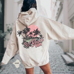 May include: Sand-coloured hoodie featuring a beach-themed graphic on the back. The design includes palm trees, a pink sunset, and the words "Beach Bum". The hoodie has a drawstring hood and long sleeves.