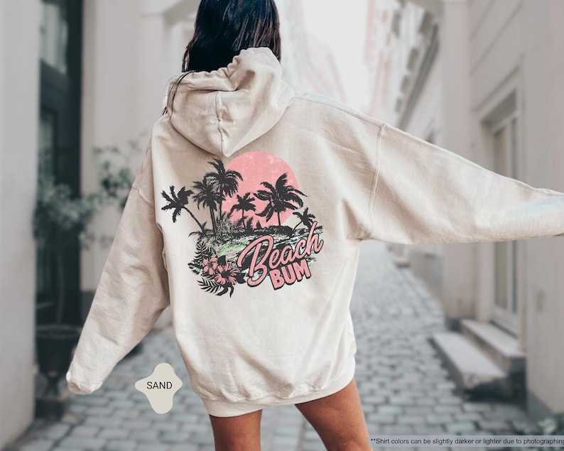 May include: Sand-coloured hoodie featuring a beach-themed graphic on the back. The design includes palm trees, a pink sunset, and the words "Beach Bum". The hoodie has a drawstring hood and long sleeves.