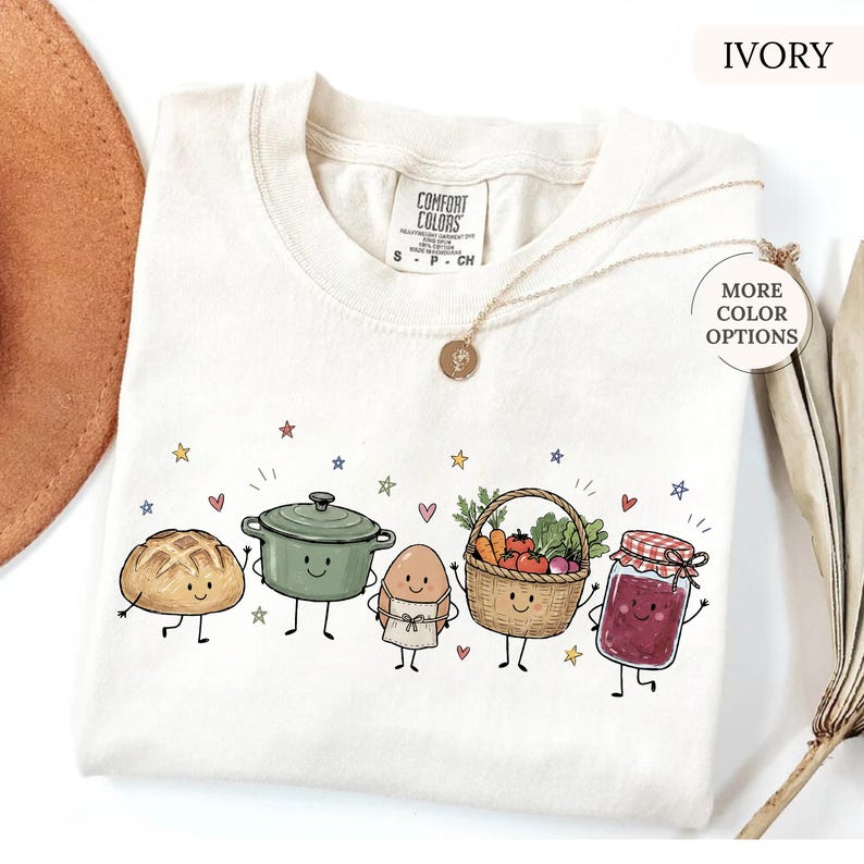 May include: An ivory t-shirt featuring a graphic print of five anthropomorphic food items: a loaf of bread, a green pot, an egg wearing an apron, a basket of vegetables, and a jar of jam. The shirt has a Comfort Colors label at the neck.