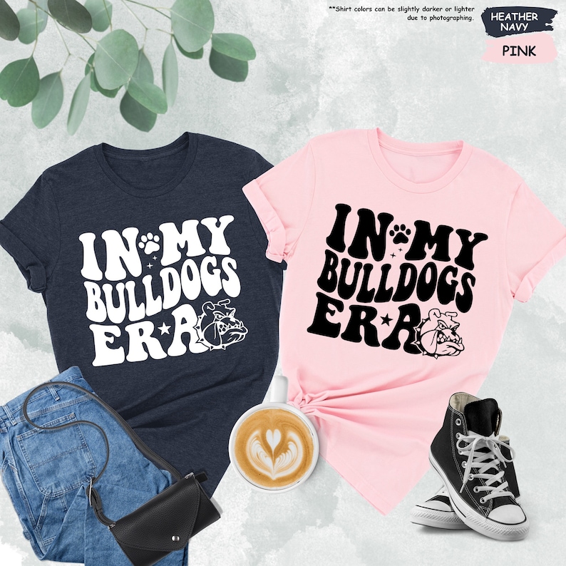 May include: Two t-shirts, one navy blue and one pink, with the text "In My Bulldogs Era" in a retro font. Each shirt features a cartoon bulldog graphic.