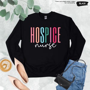 K&ouml;nnte beinhalten: Schwarzer Pullover mit dem Text "HOSPICE nurse" in Regenbogenfarben.