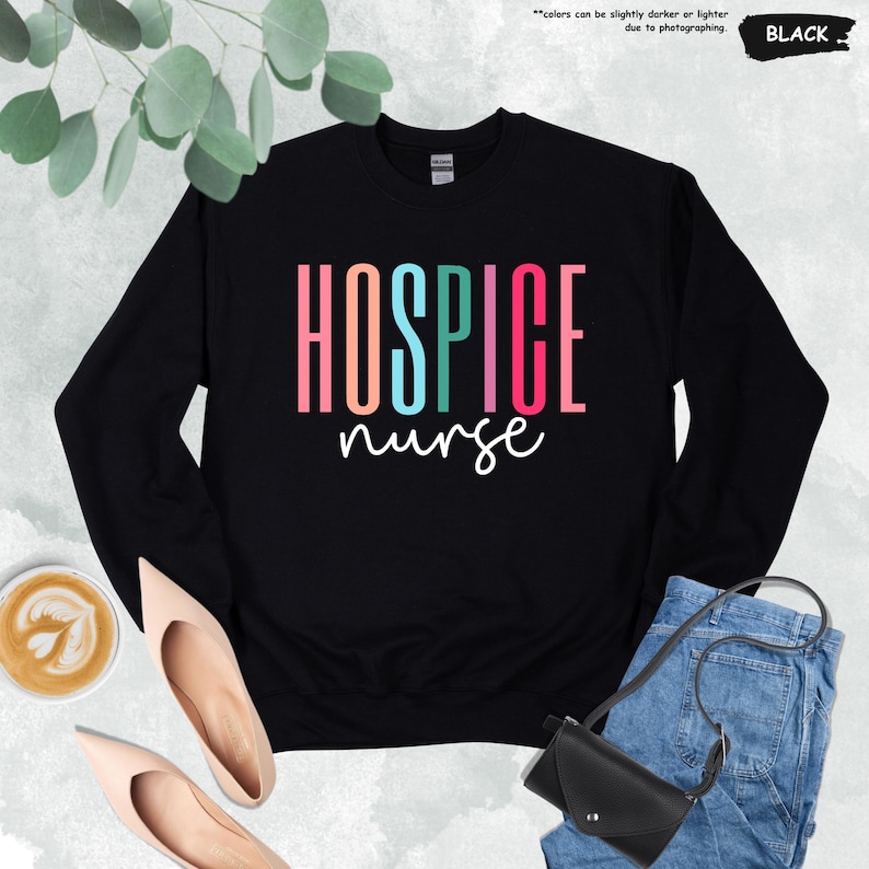 K&ouml;nnte beinhalten: Schwarzer Pullover mit dem Text "HOSPICE nurse" in Regenbogenfarben.