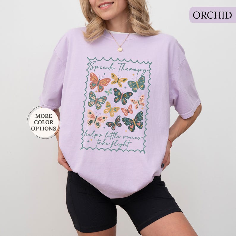 Puede incluir: Camiseta color orqu&iacute;dea con un gr&aacute;fico de mariposas dentro de un marco decorativo. El texto "Speech Therapy" y "helps little voices take flight" est&aacute;n impresos. La camiseta es de un material suave y c&oacute;modo.