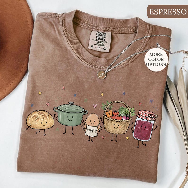 May include: An espresso-colored t-shirt featuring a graphic of five anthropomorphic food items: a loaf of bread, a green pot, an egg wearing an apron, a basket of vegetables, and a jar of jam. The shirt includes a silver necklace and text labels.