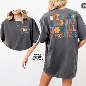 May include: Dark grey t-shirt with the text "It's a good day to fill prescriptions" on the back in colourful, retro-style lettering. The front features a smaller design with pharmacy-related graphics and the name "Sophia".