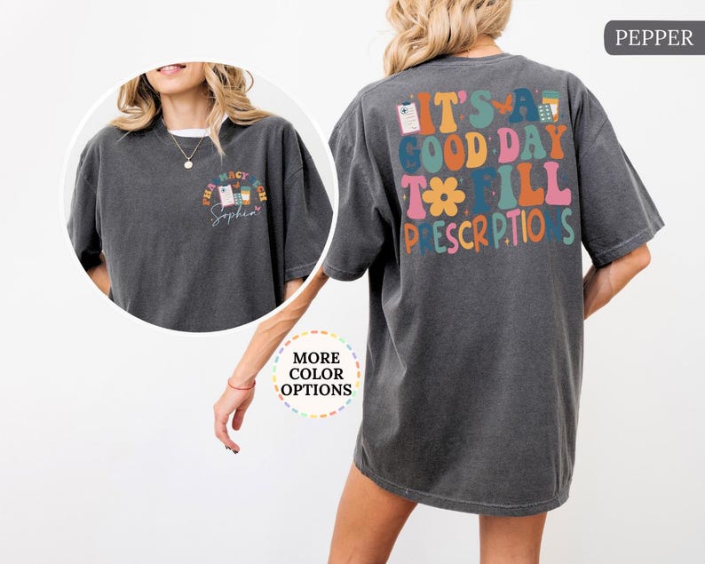 May include: Dark grey t-shirt with the text "It's a good day to fill prescriptions" on the back in colourful, retro-style lettering. The front features a smaller design with pharmacy-related graphics and the name "Sophia".