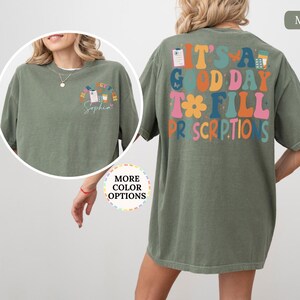 May include: A moss green t-shirt with the phrase "IT'S A GOOD DAY TO FILL PRESCRIPTIONS" in colourful, retro lettering on the back. The front has a smaller design with the text "HAPPY CV TECH" and a name. A relaxed, comfortable style.