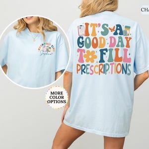 May include: Pale blue t-shirt featuring the phrase "IT'S A GOOD DAY TO FILL PRESCRIPTIONS" in various colours on the back. The front displays a smaller design with pharmacy-related graphics and the words "PHARMACY TECH".