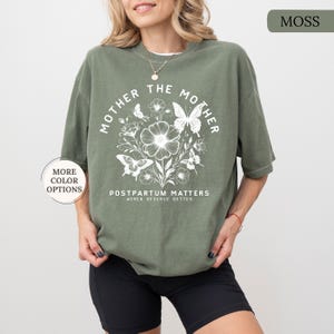 Może przedstawiać: Zielony T-shirt z białym nadrukiem graficznym, na kt&oacute;rym widnieje napis "Mother the Mother" z kwiatowym wzorem i motylami. Tekst pod grafiką brzmi "Postpartum Matters Women Deserve Better".