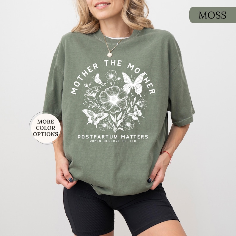 Może przedstawiać: Zielony T-shirt z białym nadrukiem graficznym, na kt&oacute;rym widnieje napis "Mother the Mother" z kwiatowym wzorem i motylami. Tekst pod grafiką brzmi "Postpartum Matters Women Deserve Better".