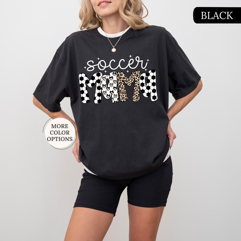 May include: Black t-shirt featuring the words "soccer mom" in a mix of patterns, including soccer balls, dog faces, and leopard print. The shirt has short sleeves and a crew neck. The words "More Color Options" are in a circle on the left.