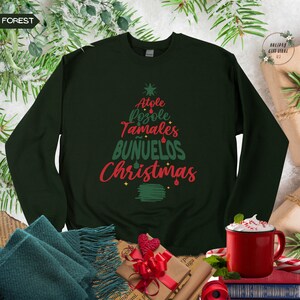 May include: A dark green sweatshirt with a Christmas tree design featuring the words "Atole", "Pozole", "Tamales", "Bu&ntilde;uelos", and "Christmas" in red and green lettering.