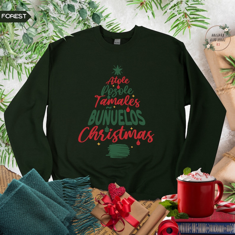 May include: A dark green sweatshirt with a Christmas tree design featuring the words "Atole", "Pozole", "Tamales", "Bu&ntilde;uelos", and "Christmas" in red and green lettering.