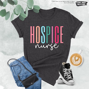 K&ouml;nnte beinhalten: Ein dunkelgraues T-Shirt mit dem Text "HOSPICE nurse" in Regenbogenfarben. Das Shirt liegt auf einer wei&szlig;en Oberfl&auml;che mit Jeans, einer Handtasche und einer Tasse Kaffee.