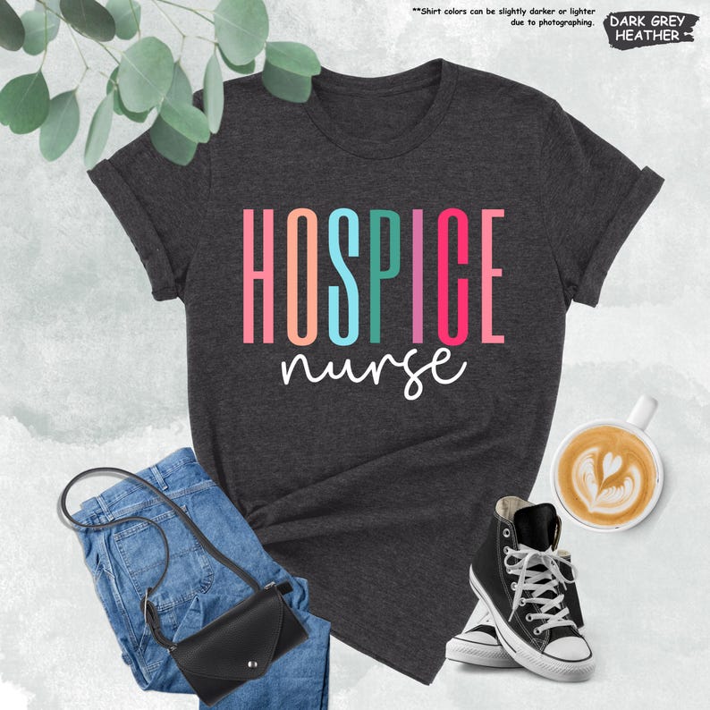 K&ouml;nnte beinhalten: Ein dunkelgraues T-Shirt mit dem Text "HOSPICE nurse" in Regenbogenfarben. Das Shirt liegt auf einer wei&szlig;en Oberfl&auml;che mit Jeans, einer Handtasche und einer Tasse Kaffee.