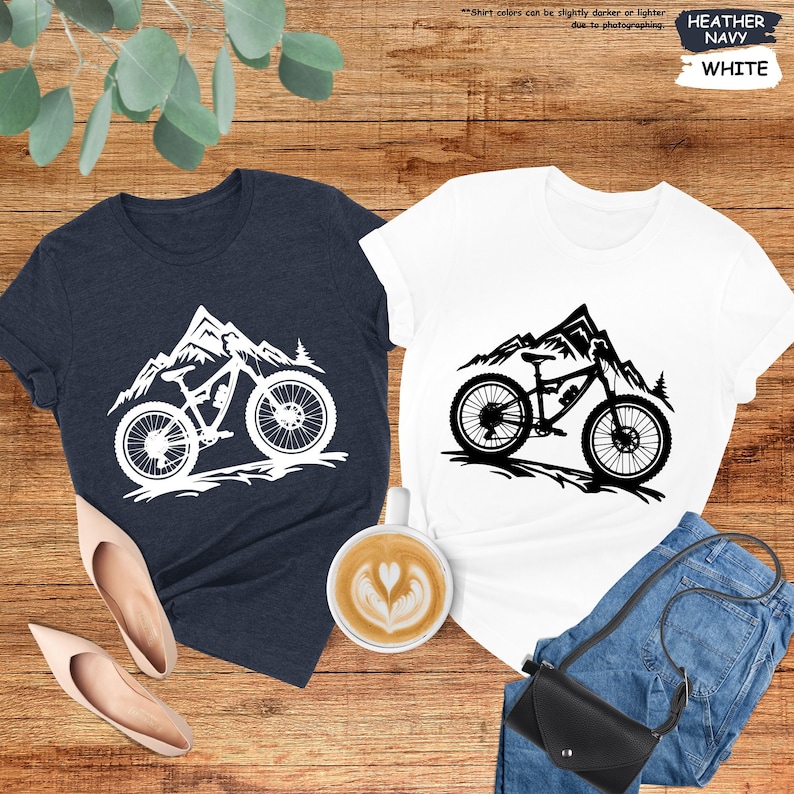 Peut inclure: Deux t-shirts avec un motif de silhouette de v&eacute;lo de montagne. Le motif repr&eacute;sente un v&eacute;lo de montagne avec une cha&icirc;ne de montagnes en arri&egrave;re-plan. Un t-shirt est blanc avec un motif noir, et l'autre est bleu marine avec un motif blanc. Le texte "Les couleurs des t-shirts peuvent &ecirc;tre l&eacute;g&egrave;rement plus fonc&eacute;es ou plus claires en raison de la photographie." est en haut de l'image. Le texte "HEATHER NAVY" et "WHITE" est en haut &agrave; droite de l'image.