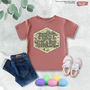 May include: A heather mauve t-shirt with "FIRST GRADE" in a camouflage design, alongside blue jeans and white trainers. Colourful Easter eggs are arranged at the bottom. The text "HEATHER MAUVE" is in the upper right corner.