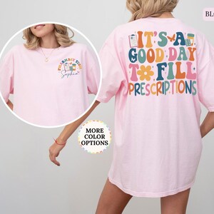 May include: A pale pink t-shirt featuring the phrase "IT'S A GOOD DAY TO FILL PRESCRIPTIONS" in vibrant colours on the back. The front displays a small graphic with "Pharmacy Tech" and a name. The t-shirt has a relaxed fit.
