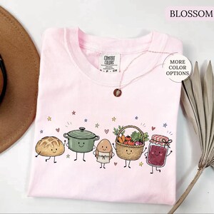 May include: A light pink t-shirt featuring a graphic print of five anthropomorphic food items: a loaf of bread, a green cooking pot, an egg wearing an apron, a basket of vegetables, and a jar of jam, all surrounded by small colorful stars and hearts.