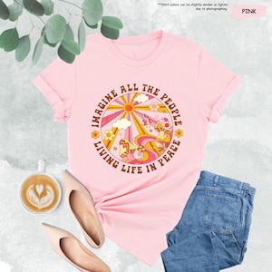 May include: Pink t-shirt with a retro-style graphic featuring a sun, clouds, mushrooms, and the text "Imagine All The People Living Life In Peace".
