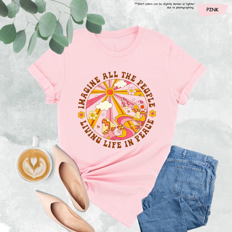 May include: Pink t-shirt with a retro-style graphic featuring a sun, clouds, mushrooms, and the text "Imagine All The People Living Life In Peace".