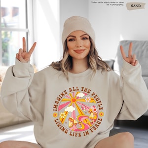 May include: A beige sweatshirt with a retro-style graphic print of a sun, clouds, flowers, and mushrooms. The text "Imagine All The People Living Life In Peace" is printed around the graphic.
