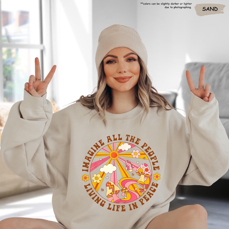 May include: A beige sweatshirt with a retro-style graphic print of a sun, clouds, flowers, and mushrooms. The text "Imagine All The People Living Life In Peace" is printed around the graphic.