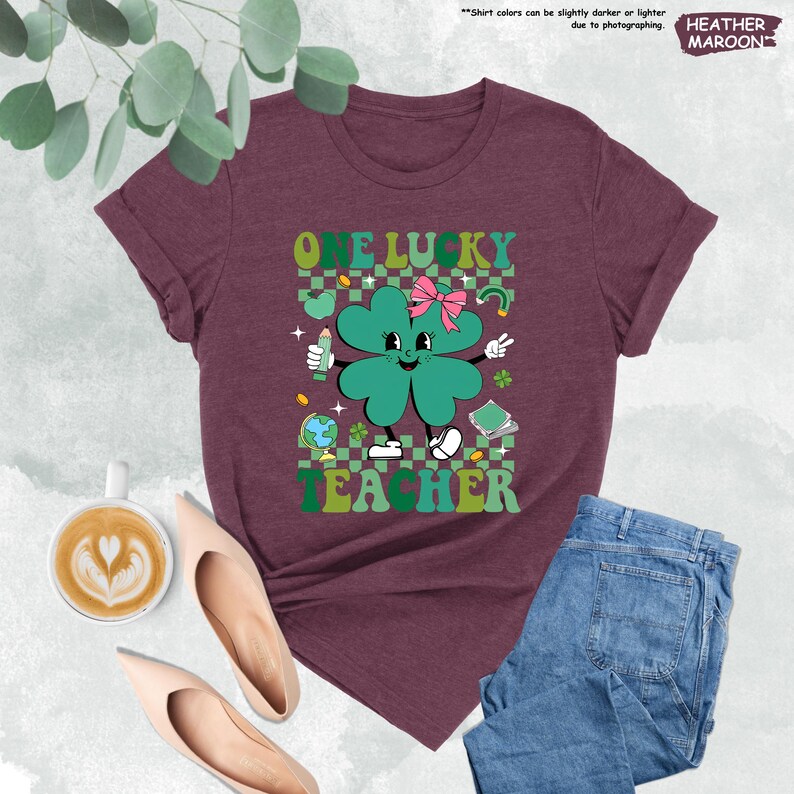 May include: A heather maroon t-shirt with a green four-leaf clover graphic. The clover has a pink bow and a smiling face. The text "One Lucky Teacher" is printed below the clover.