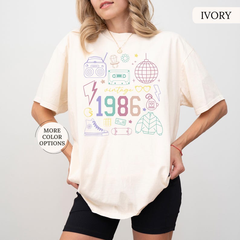 May include: Ivory-colored t-shirt with a colorful 1986 graphic. The design includes a boombox, cassette tape, disco ball, and other vintage elements. The shirt has a relaxed fit and short sleeves.