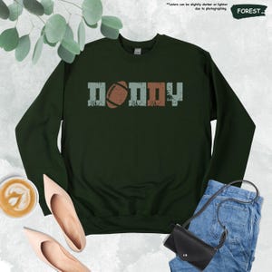 May include: A dark green sweatshirt with the word "DADDY" printed in a vintage style with a football graphic in the middle.