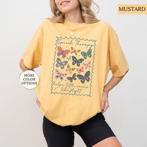 Puede incluir: Camiseta de color mostaza con un dise&ntilde;o rectangular con mariposas y el texto "Speech Therapy helps little voices take flight." El dise&ntilde;o incluye mariposas de varios colores. La camiseta tiene mangas cortas.