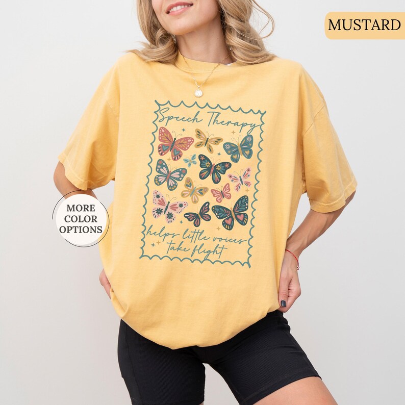 Puede incluir: Camiseta de color mostaza con un dise&ntilde;o rectangular con mariposas y el texto "Speech Therapy helps little voices take flight." El dise&ntilde;o incluye mariposas de varios colores. La camiseta tiene mangas cortas.