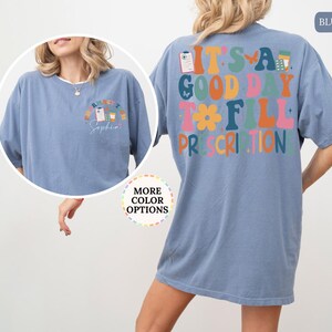 May include: Blue jean coloured t-shirt with the text "It's a good day to fill prescriptions" on the back. The front of the shirt has a smaller design with the text "Pharmacist Sophia". The shirt has short sleeves and a crew neck.